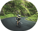 A cyclist seen from behind riding on a winding road through a vibrant green, mountainous landscape.