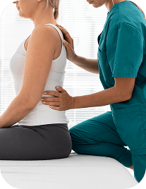 A female physical therapist in green scrubs performs manual therapy on a female patient's back as she sits on a treatment table.
